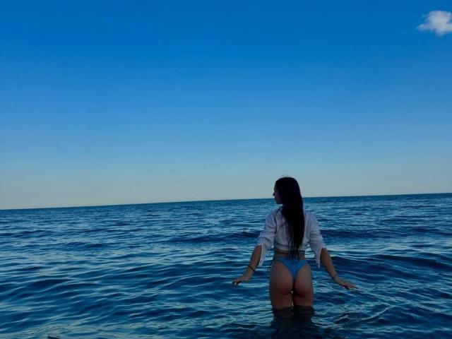PaigePrado with long dark hair, white shirt, and blue thong standing in clear ocean water, facing away, showing her ass and back, under a bright blue sky.