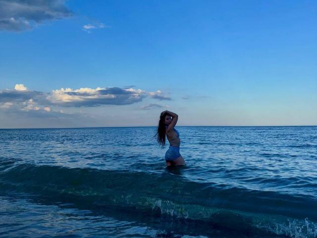 PaigePrado stands waist-deep in ocean water, back to camera, arms raised, wearing a blue bikini, her ass and tits visible, long brown hair, clear blue sky above.