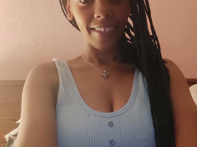 MelissaCrush with dark skin, braided hair, smiling, wearing a light blue ribbed tank top with buttons, showing small tits, necklace with a heart pendant.