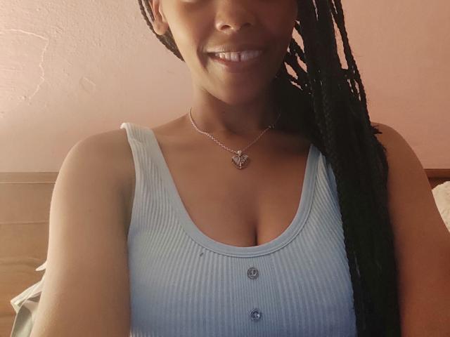 MelissaCrush with dark skin, smiling, wearing a light blue ribbed tank top, buttoned down, showing her tits, long black braids, silver necklace with butterfly pendant.