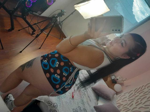 CharlottexEvans with long black hair in a ponytail, white tank top, blue Cookie Monster shorts, tattoo on thigh, holding phone, winking, standing in room with wooden floor.