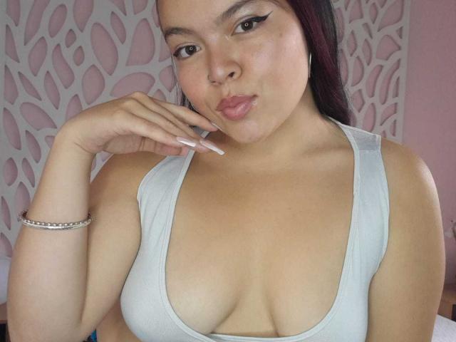 CharlottexEvans with pouty lips, pale skin, dark hair, white tank top, showing large tits, right hand near mouth, long nails, silver bracelet, looking at camera.