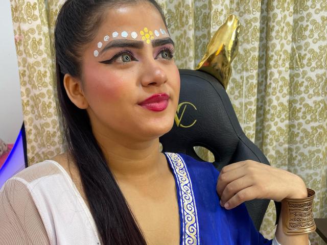 NayaYalitza has brown skin, black hair in a ponytail, wearing a blue sari with white trim, gold headpiece, white face dots, red lipstick, and gold bangles, smiling.