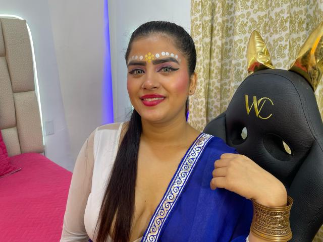 NayaYalitza with dark hair in a ponytail, wearing a blue sari, white forehead dot, gold mask with "W", red lipstick, smiling, holding mask, gold bangles, pink bed.