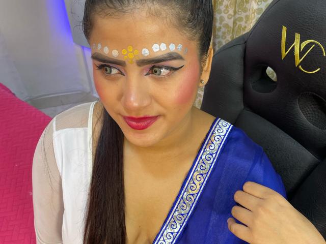 NayaYalitza with dark hair in a ponytail, wearing a blue sari with white pattern, red lipstick, and white makeup dots on her forehead, smiling at the camera. Boobs visible.