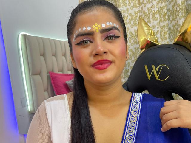NayaYalitza has dark hair, brown eyes, pink eyelids, white and gold face dots, red lipstick, blue and white dress, holding a black W-shaped headpiece with gold ears.