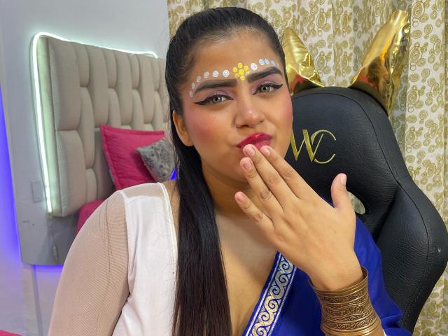 NayaYalitza with dark hair in a ponytail, wearing a blue and white dress, gold bangles, and a floral face mask, kissing her finger with pink lipstick and bold eye makeup.