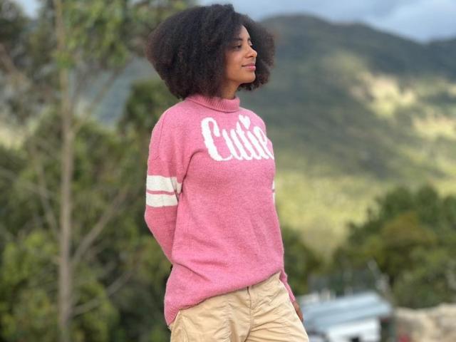 EmmyRacheel with curly black hair, pink sweater, white stripes, and beige pants, standing with hands in pockets, smiling, outdoors with green hills in the background.