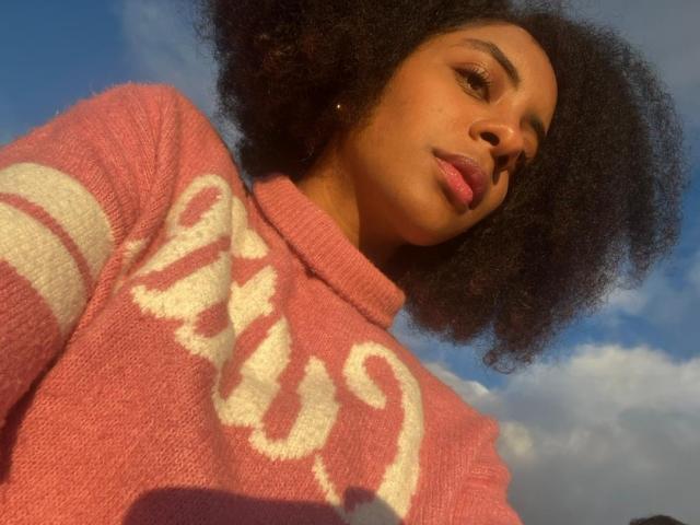 EmmyRacheel with dark, fluffy afro wearing a pink sweater with white lettering, looking down with a subtle smile, clear blue sky in background.