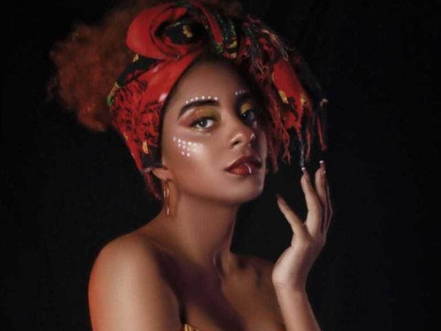 EmmyRacheel with red headwrap, yellow-green eye makeup, white facial markings, red lips, gold earrings, touching her face, dark background, bare shoulders, looking up with seductive gaze.