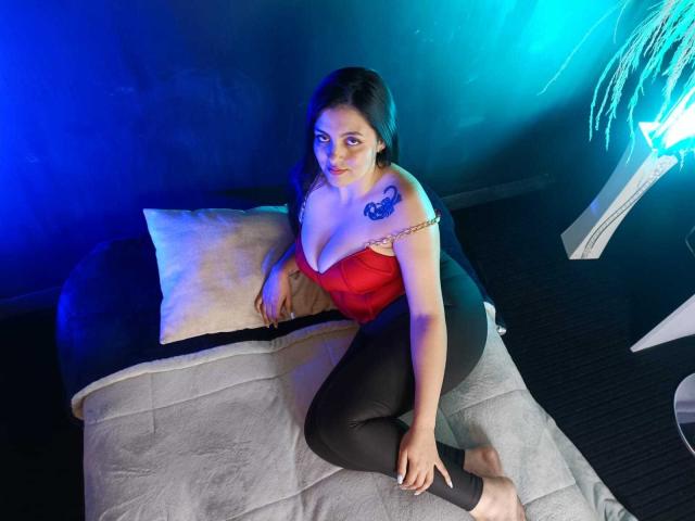 AntonellaRussel, with big tits, red top, and black pants, sits on a bed with a pillow, blue and purple lights, and a plant in the background. She has a tattoo.