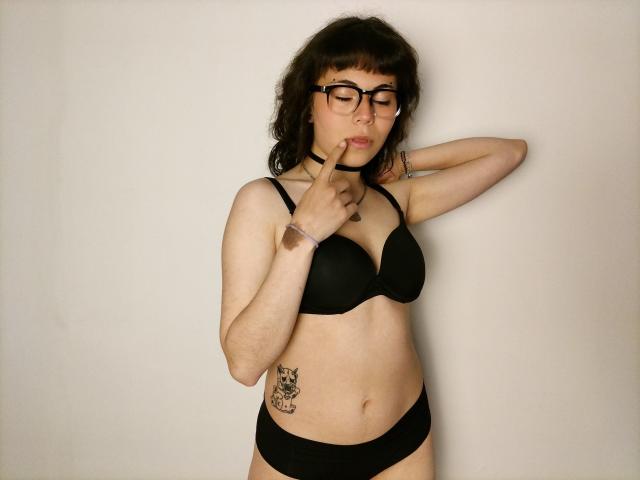BeautyInsomnia with glasses, black bra, and panties, finger to lips, tattoo on left side, choker, brunette, fair skin, slight belly, black hair, standing, arm raised.
