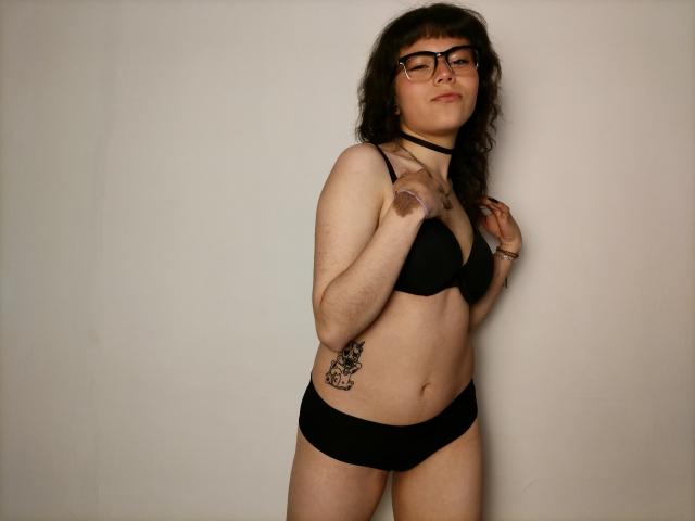 BeautyInsomnia with curly dark hair, glasses, black choker, black bra, and black panties, standing, hand on chest, tattoo on right side, confident, subtle smile, white background.