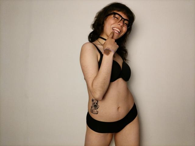 BeautyInsomnia, with glasses, black bra, and panties, has a tattoo on her right hip, smiling, and touching her chin with a finger, showing a playful, confident expression.