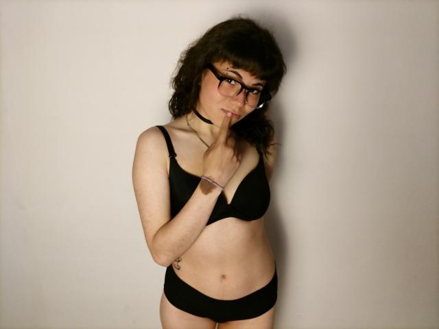 BeautyInsomnia with curly brown hair, glasses, black choker, and black bra and panties, biting her finger, showing a tattoo on her left wrist and small belly button.