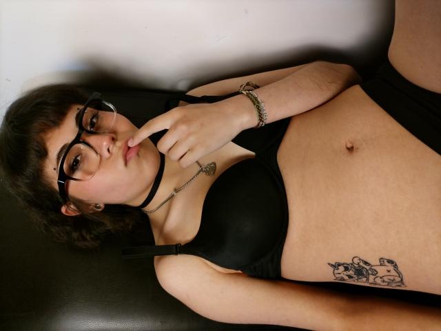 BeautyInsomnia with glasses, black bra, and choker, finger on lips, showing tits, belly tattoo, and black underwear, lying on dark leather. She's cute, with a silver bracelet.