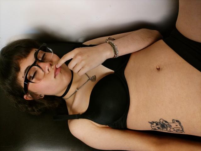 BeautyInsomnia with glasses, black bra, and choker, biting finger, showing tattoo on right side, lying on black surface, wearing bracelet, with short dark hair, and light skin.