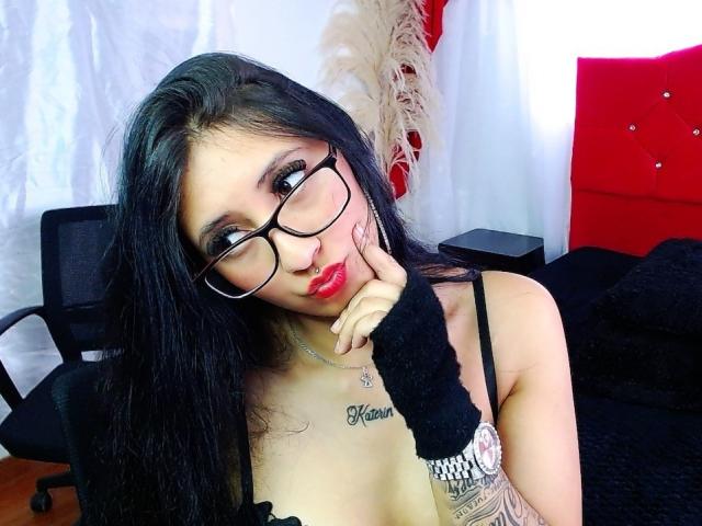 WendyD con cabello negro, gafas, lápiz labial rojo, sujetador de encaje negro, mano en la mejilla, tatuaje debajo del mentón, sosteniendo un calcetín negro, sentada en una silla negra, cabecera roja en el fondo..