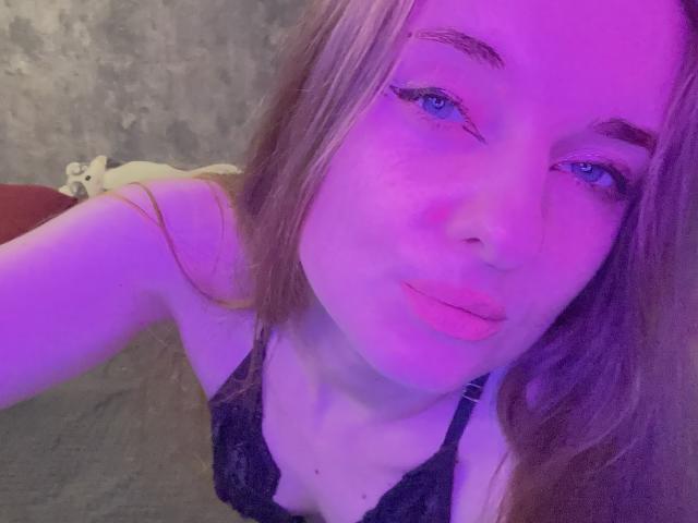 KariMillie with pouty pink lips, blue eyes, and long brown hair, wearing black lace lingerie, has a purple-hued face and looks directly at the camera.