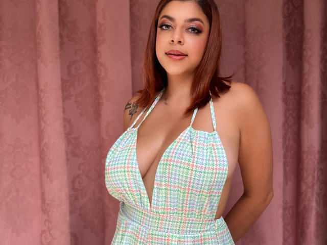 MedeaHelios with big boobs in a colorful checkered halter dress, brown hair, tattoo on right shoulder, pink lips, smiling, standing against pink curtain, hands behind back.