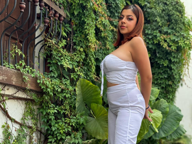 MedeaHelios has big tits in a white strapless top and white pants, standing against a wall covered in green vines, with brown hair and a slight smile.