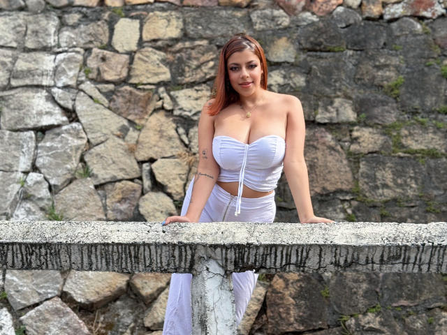 MedeaHelios with big tits in a white strapless top and white pants, leaning on a stone wall, has long red hair and a tattoo on her left arm.