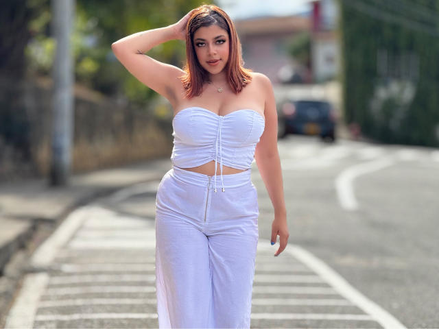 MedeaHelios with red hair, white strapless top showing big tits, and white pants, standing on a sidewalk, hand in hair, looking at camera, curvy, sexy.