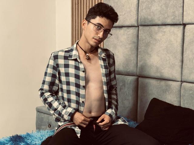 AnryuOswald with glasses, short curly black hair, open checkered shirt, no pants, sitting on blue fuzzy bed, black pants around thighs, necklace, exposed chest, looking at camera.