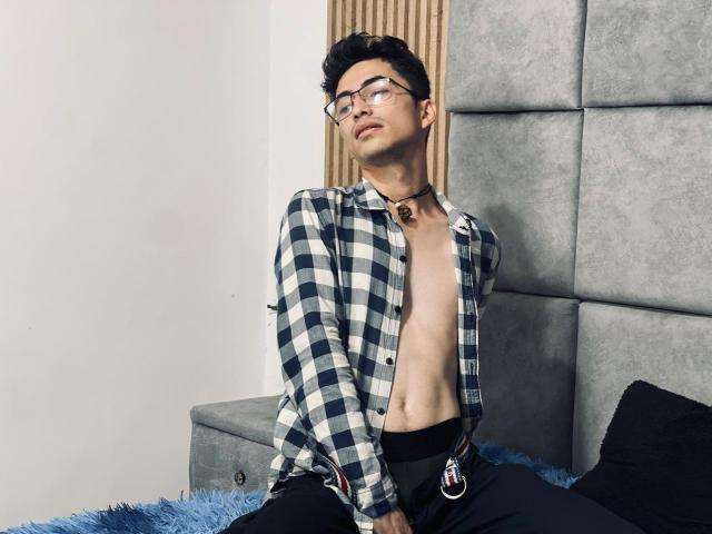 AnryuOswald, shirtless, wearing glasses, checkered shirt, black pants, collar with bell, sitting on bed, blue blanket, grey headboard, looking down, short black hair, slight smile.