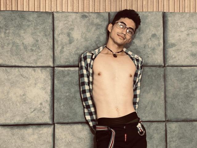 AnryuOswald, shirtless, wearing glasses, black necklace, black pants, hands in pockets, leaning against grey wall, short dark hair, slight smile, chest visible, flat abs.