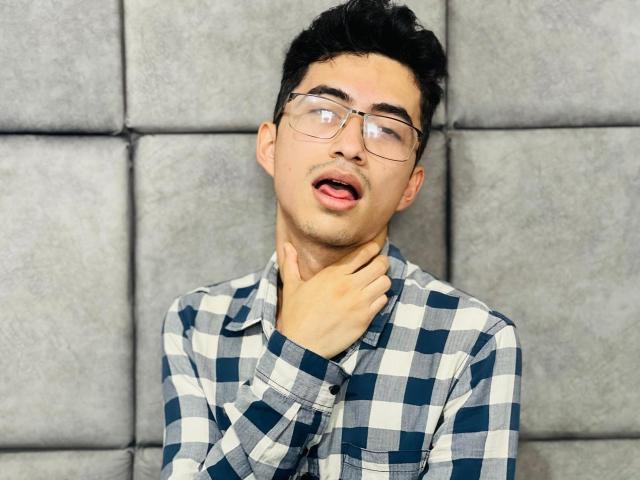 AnryuOswald with glasses, black hair, and a slightly open mouth, gripping his neck with his right hand, wearing a blue and white checkered shirt.