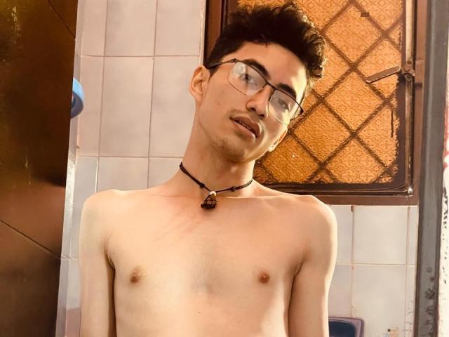 AnryuOswald, shirtless, with glasses, short dark hair, choker with a black stone, small tits, pale skin, leaning slightly to the side, looking up, in a dimly lit room.