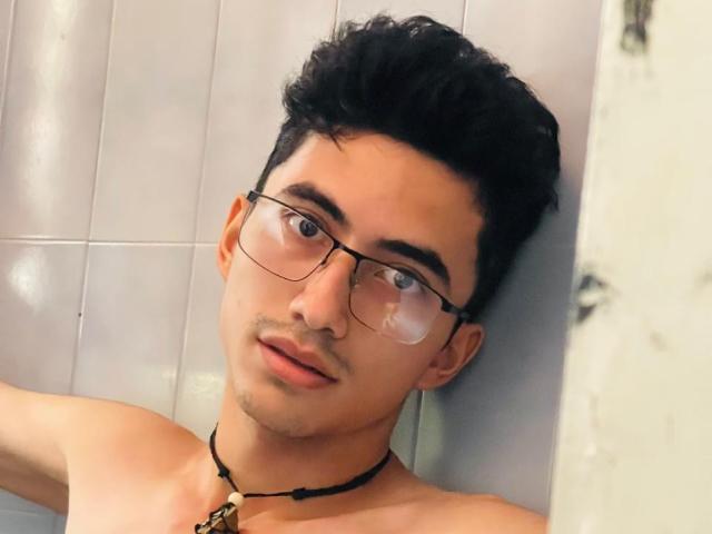 AnryuOswald with dark curly hair, glasses, and a necklace, leaning against a wall, looking at the camera, shirtless, with a slight smirk and visible collarbone.