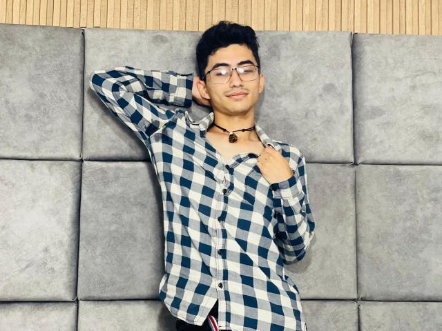 AnryuOswald with glasses, black choker, checkered shirt, hand behind head, slight smile, short black hair, medium skin, standing against gray wall.