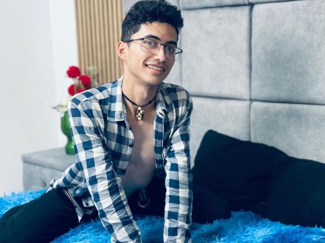 AnryuOswald, wearing glasses and a blue check shirt, smiling with a chest necklace, sitting on a blue furry bed, black pants, and black pillows in the background.