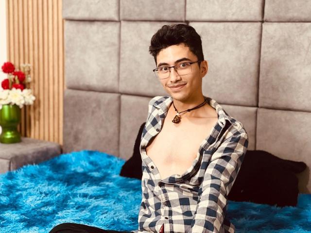 AnryuOswald, wearing glasses and a partially unbuttoned checkered shirt, exposing his chest, sits on a blue fuzzy bed, smiling, with a necklace.