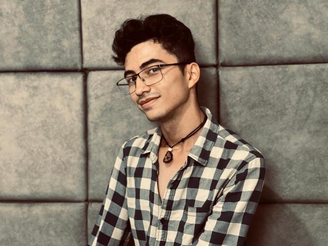 AnryuOswald with curly black hair, glasses, and a slight smirk, wearing a black-and-white checkered shirt, and a black necklace with a round pendant.