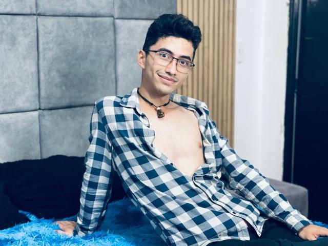 AnryuOswald with glasses, black hair, and a cute smile, wearing an open checkered shirt, revealing a bare chest, sitting on a blue fuzzy bed, necklace with a pendant.