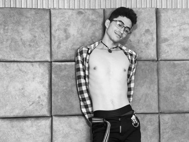 AnryuOswald, shirtless, with glasses, a checkered open shirt, and dark pants, leaning against a padded wall, smiling, showing a flat chest and a small, cute belly.