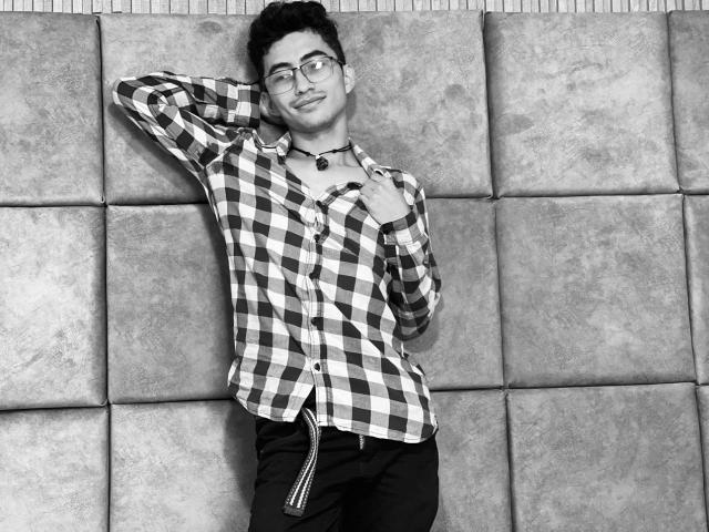 AnryuOswald with glasses, checking shirt buttons, hand behind head, plaid shirt, black pants, necklace, standing against padded wall, smiling, short dark hair, medium build, black-and-white photo.