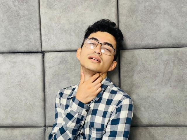 AnryuOswald with glasses, black hair, and a checkered shirt, has eyes closed and a hand on his neck, looking slightly annoyed. He's against a gray, padded wall.