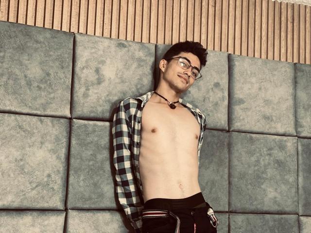 AnryuOswald, shirtless, with glasses, wearing an open plaid shirt, black pants, and a necklace, standing against a grey wall, showing a slim, flat-chested, hairy chest.