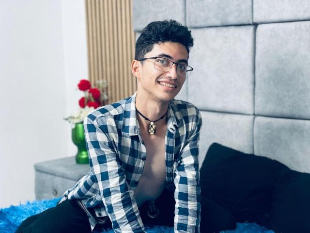AnryuOswald, shirtless with a checkered blue and white shirt, black pants, glasses, necklace, smiling, dark curly hair, sitting on a blue bed, black pillow in background.