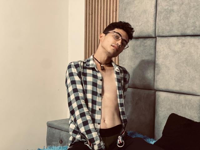 AnryuOswald, shirtless with blue and white checkered flannel, black pants, and glasses, leaning back on bed, looking down, necklace, semi-erect, short dark hair, soft lighting, relaxed.