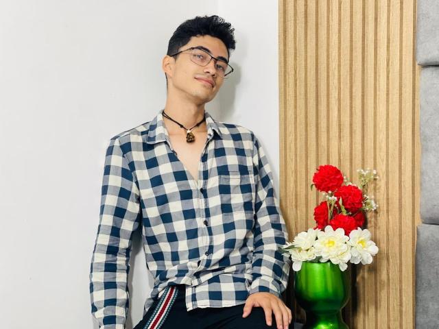AnryuOswald, wearing glasses, a blue and white checkered shirt, and a black necklace, leans against a wooden wall with a green vase of red and white flowers.