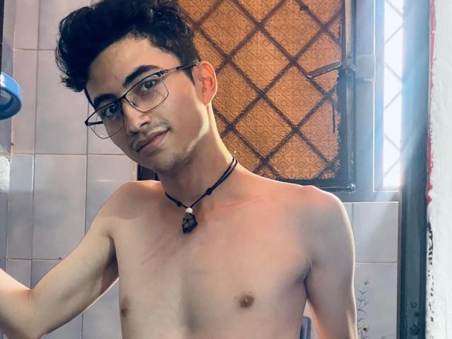AnryuOswald is shirtless with curly black hair, glasses, and a black necklace with a stone, standing in a bathroom with a diamond-patterned window. He's looking at the camera.