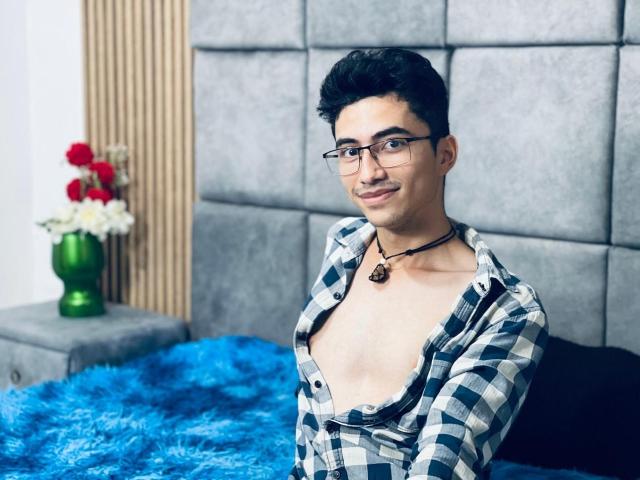 AnryuOswald with glasses, black curly hair, open checkered shirt, exposing chest, black necklace, sitting on blue fuzzy bed, smiling. Background: gray padded wall, green vase with flowers.