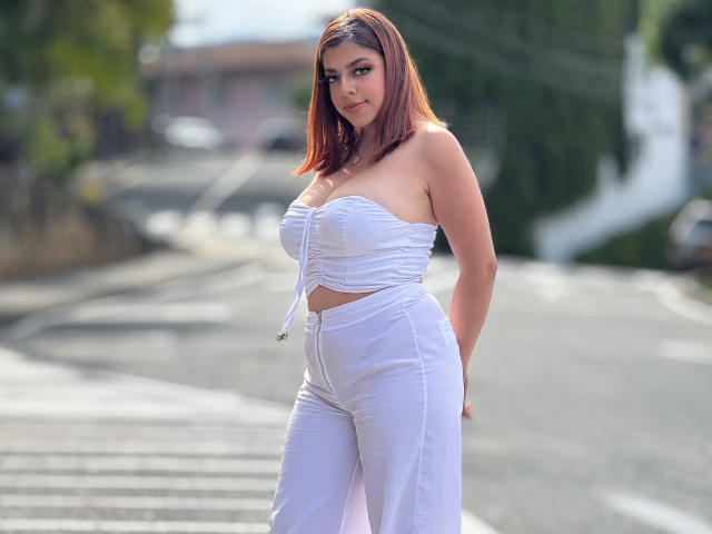 MedeaHelios with red hair, white strapless top showing her boobs, and white pants standing on a sunlit street, hands behind her back, looking at the camera.