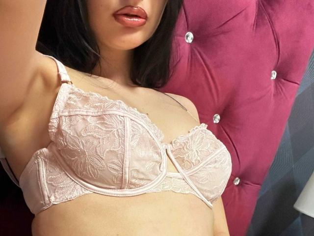 ScarlettEve wearing a pink lace bra with floral patterns, showing her tits and slightly parted lips with glossy pink lipstick. Her arm is raised, taking a selfie.