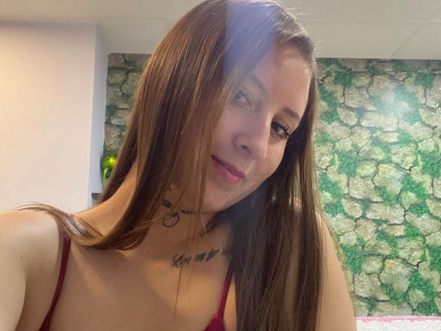 EvaDaMore with long brown hair, red spaghetti strap top, tattoo below neck, slight smile, subtle light in eyes, and casual makeup. Her tits are small, she's cute.
